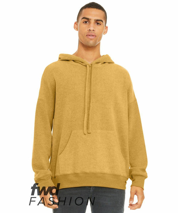 A young person wearing a mustard-colored Bella Canvas BE130 hoodie with a front pocket and drawstrings stands against a white background. They have a neutral expression and their hands are by their sides. The text "fwd FASHION" is displayed on the lower part of the image.