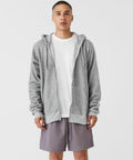 A person stands facing forward, wearing a plain white Bella Canvas BE131 t-shirt under an open gray zip-up hoodie. They have on light purple shorts. The background is a plain white.