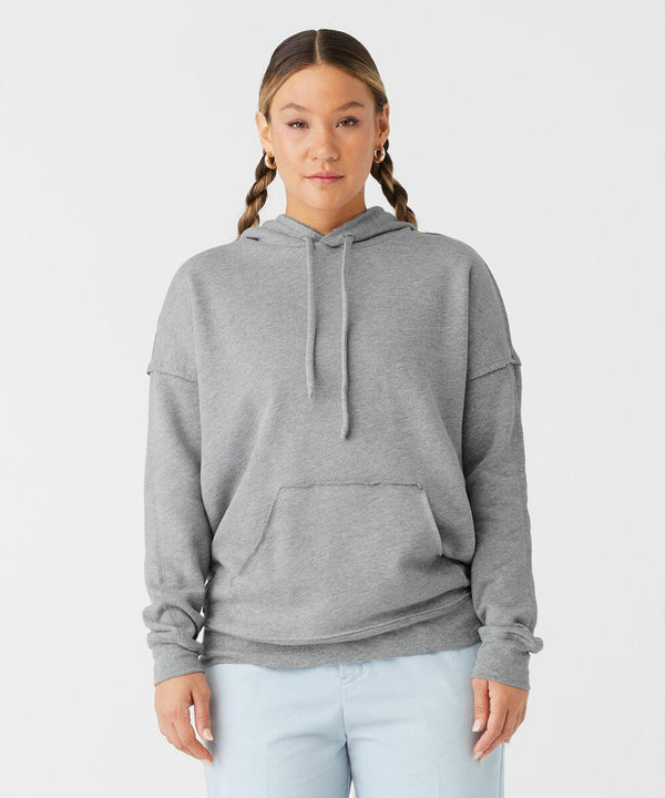 A person with braided hair stands against a white background, wearing a gray Bella Canvas BE132 hooded sweatshirt with the hood down and their hands in the front pocket. They are also wearing light blue pants and gold hoop earrings, looking directly at the camera with a neutral expression.
