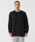 A person stands against a plain white background, wearing a black long-sleeve sweatshirt (BE133 by Bella Canvas) and light grey pants. The sweatshirt has a minimalistic design with a slight horizontal seam across the chest. The individual has a neutral expression and short hair.