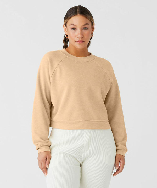 Woman stands against a plain background, wearing a BE134 long-sleeve sweatshirt from Bella Canvas and white pleated pants. She has light brown hair pulled back, and is accessorized with hoop earrings. She gazes directly at the camera with a neutral expression.