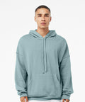 A young man with short hair wearing a Bella Canvas BE136 light blue hoodie stands against a plain white background. The hoodie has a front pocket and drawstrings, and the man has a neutral expression on his face.