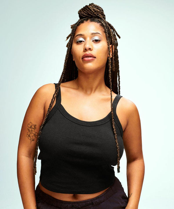 A person with long braided hair and a floral tattoo on their arm wears a black Bella Canvas BE139 tank top. They stand against a plain light background.