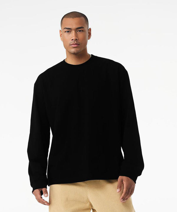 A person wearing the Bella Canvas Unisex long sleeve heavy tee in black stands against a plain white background, paired with beige trousers, looking directly at the camera with a neutral expression.
