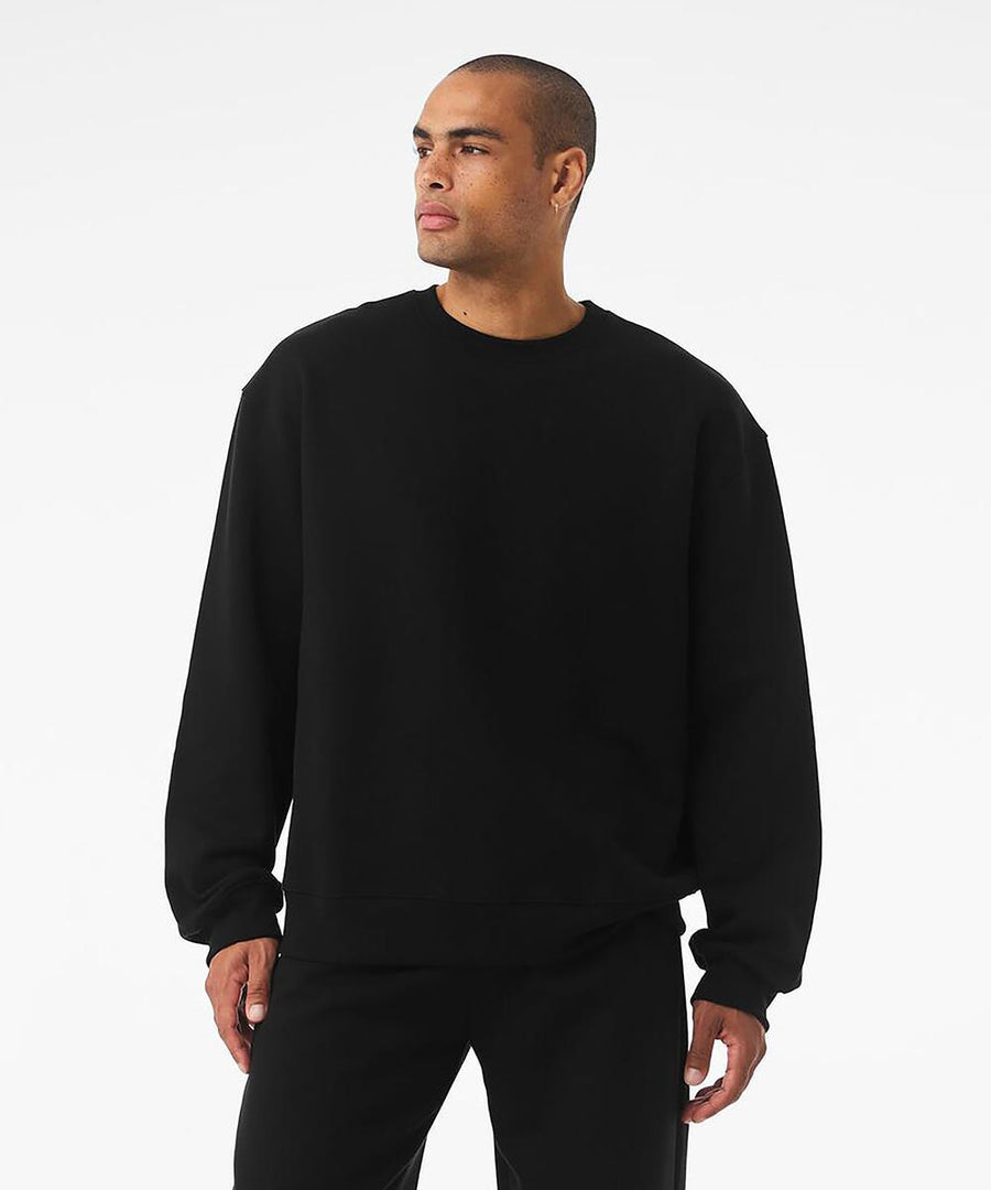 A person wears a Bella Canvas Unisex crew neck heavy sweatshirt paired with black trousers, standing against a white background and gazing to the side with a neutral expression.