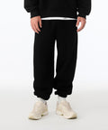 A person wears Bella Canvas Unisex heavy joggers in black with a black sweatshirt, white shirt underneath, and light trainers, standing against a plain white background. Only their lower half is visible.
