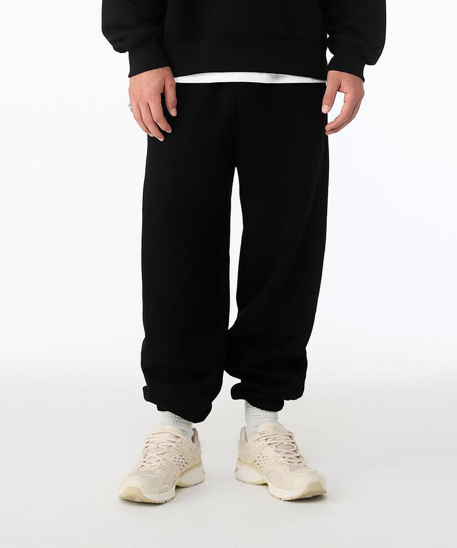 A person wears Bella Canvas Unisex heavy joggers in black with a black sweatshirt, white shirt underneath, and light trainers, standing against a plain white background. Only their lower half is visible.