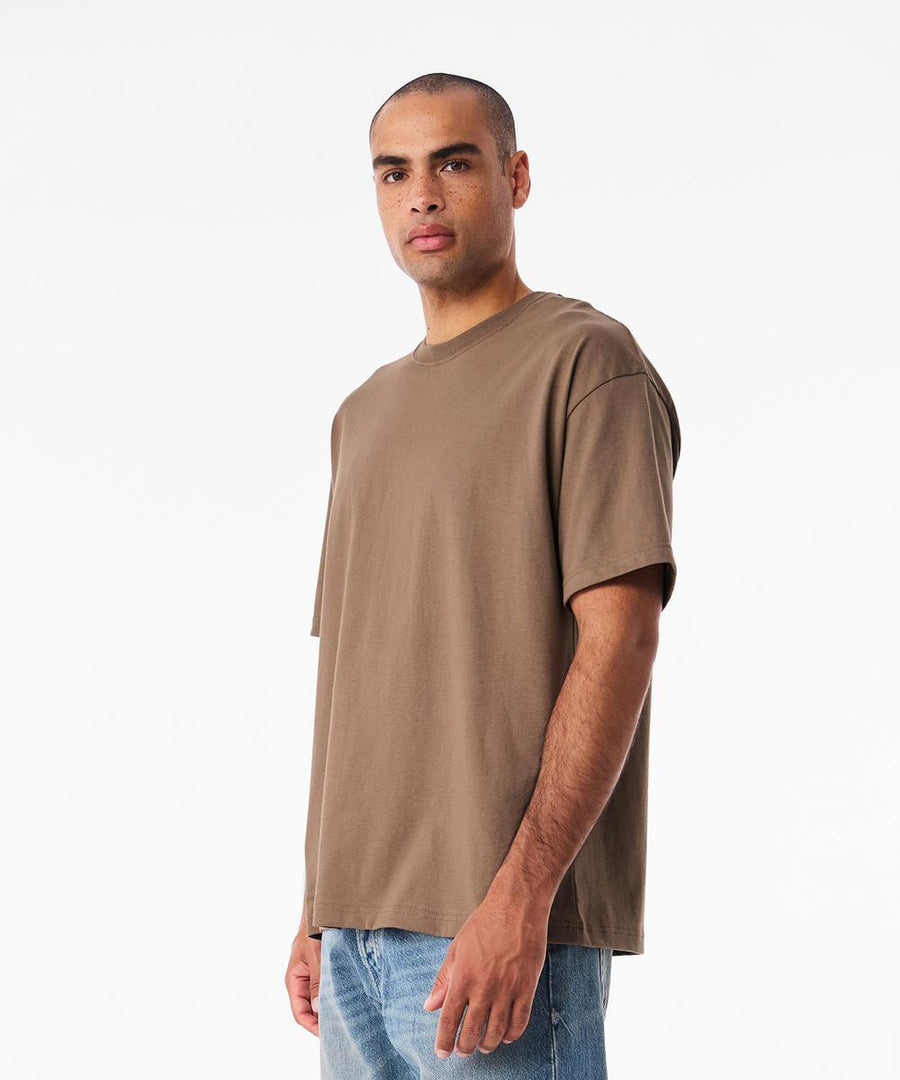 A man with short hair and a neutral expression stands slightly to the left against a plain white background, wearing a loose-fitting Bella Canvas Unisex Heavy Boxy T-shirt in brown and blue jeans.