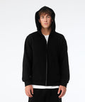 A young man faces forwards in a Bella Canvas Unisex heavy full-zip hoodie, black with the hood up, layered over a white shirt. He has a neutral expression and stands against a plain white background.