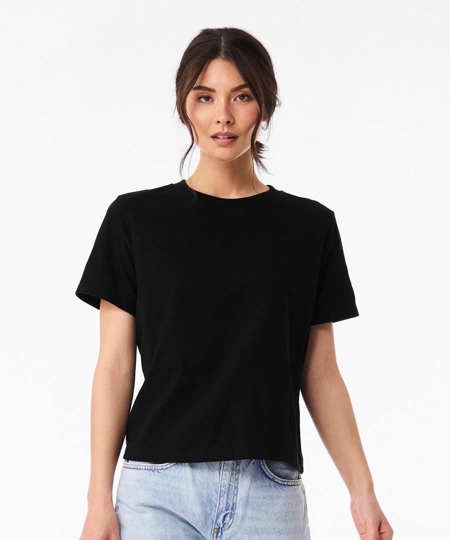 A woman with long brown hair wears a black Bella Canvas Women's heavy boxy tee and light blue jeans, standing facing forwards against a plain white background.