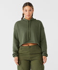 A woman stands against a plain white background, wearing an olive green cropped hoodie with long sleeves and a drawstring hood. She has her hair pulled back and is also wearing high-waisted, olive green cargo pants with multiple pockets. The hoodie is the BE220 by Bella Canvas.