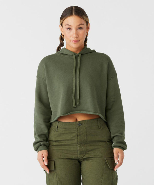 A woman stands against a plain white background, wearing an olive green cropped hoodie with long sleeves and a drawstring hood. She has her hair pulled back and is also wearing high-waisted, olive green cargo pants with multiple pockets. The hoodie is the BE220 by Bella Canvas.