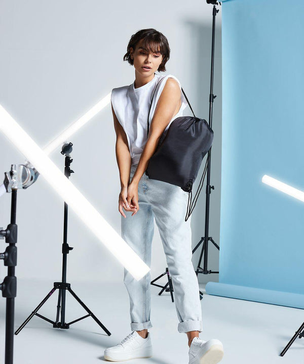 A person stands in a studio with lighting equipment, wearing a sleeveless white top, light jeans, white sneakers, and carrying a black Bagbase BG005 drawstring bag. The background includes a blue backdrop.