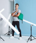 A person stands with a Bagbase BG010 slung over their shoulder in a photography studio, surrounded by studio lights and a light blue backdrop.