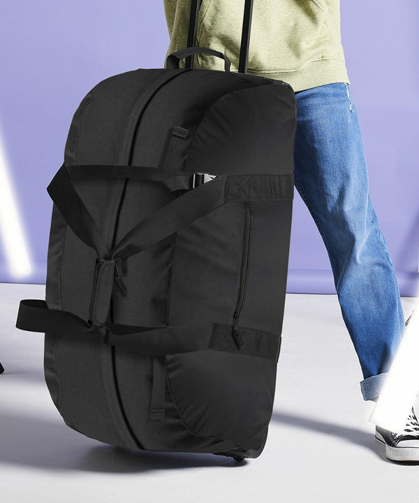 A person wearing blue jeans and a light green sweater is pulling a large black Bagbase BG023 with handles and straps. The background is purple with white light streaks. The scene appears to be a travel or airport setting.