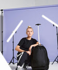 Person in black outfit crouching with a Bagbase BG025 in a studio setting with purple backdrop, light stands, and LED lights.