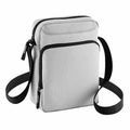 The BG030 by Bagbase is a compact, light gray fabric crossbody bag featuring black zippers and an adjustable black strap.