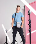 A person wearing a light blue T-shirt and black pants stands with hands in pockets, carrying a Bagbase BG030 crossbody bag, posed in front of a pink backdrop with studio lights arranged around.