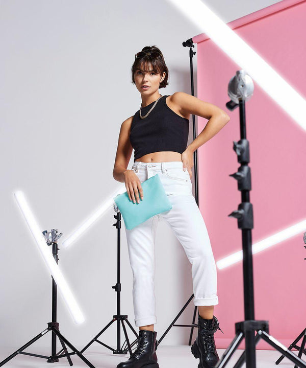 Woman posing in a studio with bright white and pink background, wearing a black tank top, white pants, black boots, and holding a turquoise BG038 by Bagbase. She stands among lighting equipment.