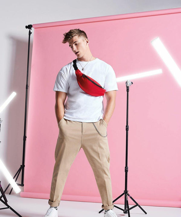 A person stands confidently in front of a pink background with white lights, wearing a white t-shirt, beige pants, white sneakers, and a Bagbase BG042 across the chest. Hands are in pockets.