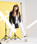 A woman with long hair stands in front of a yellow background. She is wearing a black jacket over a gray hoodie, white pants, and BG047 Bagbase yellow sneakers. There are white light beams and light stands around her.