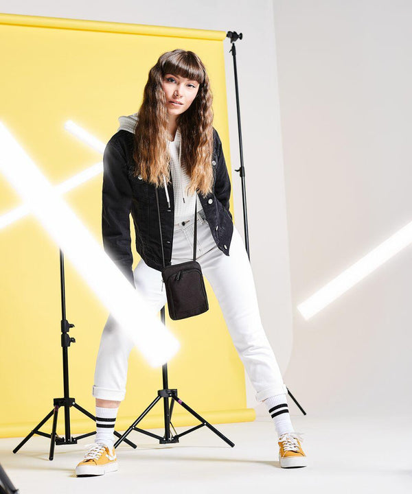 A woman with long hair stands in front of a yellow background. She is wearing a black jacket over a gray hoodie, white pants, and BG047 Bagbase yellow sneakers. There are white light beams and light stands around her.