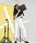 A person with long hair wearing sunglasses stands confidently in a studio with a bright yellow backdrop. They hold a black BG067 laptop sleeve by Bagbase and wear a black t-shirt, white joggers, black shoes, and yellow socks. Neon light fixtures are positioned around them.