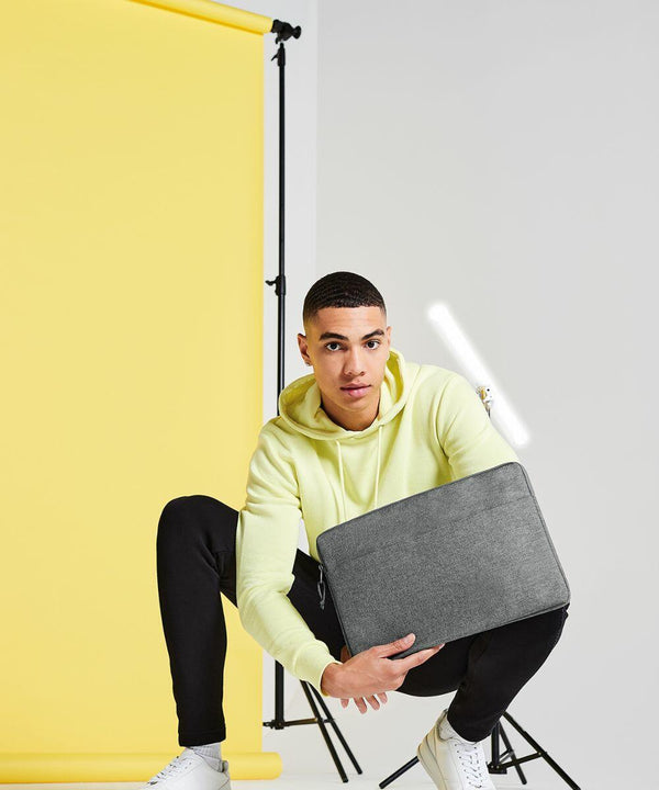 A person in a yellow hoodie and black pants squats in front of a yellow backdrop, holding a gray Bagbase BG068. The individual is wearing white sneakers and is looking at the camera. There is a white strip light in the background.