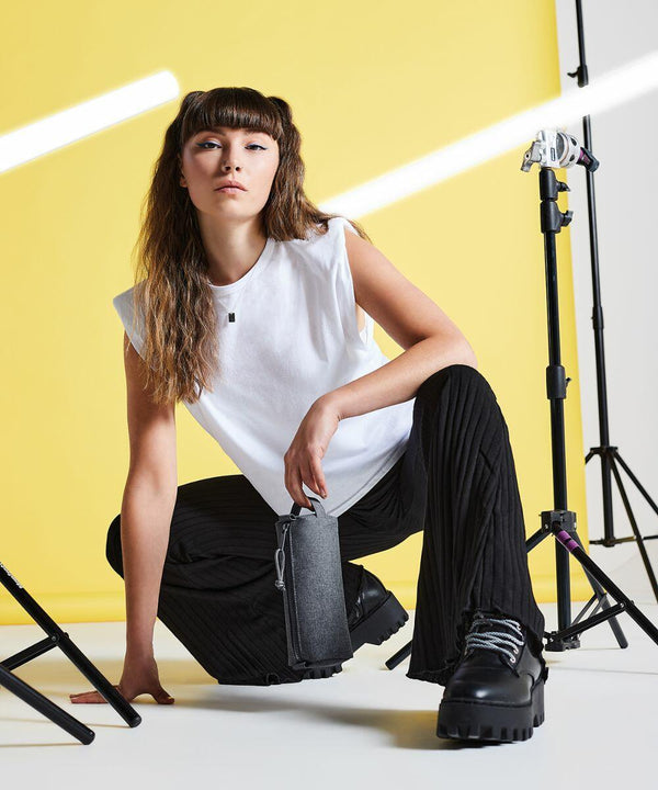 A woman with long brown hair, wearing a white sleeveless shirt and black pants, kneels on the floor holding a Bagbase BG069. She is surrounded by camera tripod stands, set against a bright yellow backdrop.
