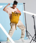 A person poses with a Bagbase BG118 mustard yellow backpack, wearing a peach and beige color-blocked shirt and beige cargo pants. The backdrop is light blue, and there are LED strip lights and tripods positioned around them, creating a dynamic studio setting.