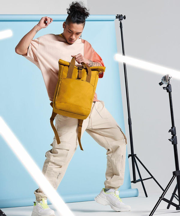 A person poses with a Bagbase BG118 mustard yellow backpack, wearing a peach and beige color-blocked shirt and beige cargo pants. The backdrop is light blue, and there are LED strip lights and tripods positioned around them, creating a dynamic studio setting.