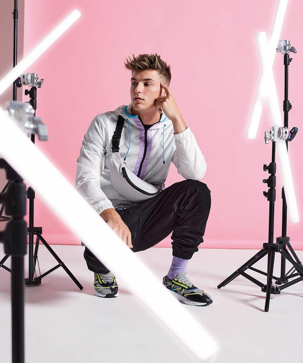 A young person wearing a white jacket with purple and light blue accents, black pants, and green sneakers crouches thoughtfully in a studio with a pink backdrop. Fluorescent light tubes are positioned around them, creating an energetic and modern vibe. They have the BG134 by Bagbase slung over their shoulder, completing their stylish look.