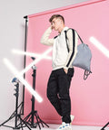 A person stands in a studio with a pink background, leaning on one leg. They wear a white and beige hoodie, black pants, and white sneakers, carrying a gray Bagbase BG137 over one shoulder. Several bright white lights and light stands are positioned around them.