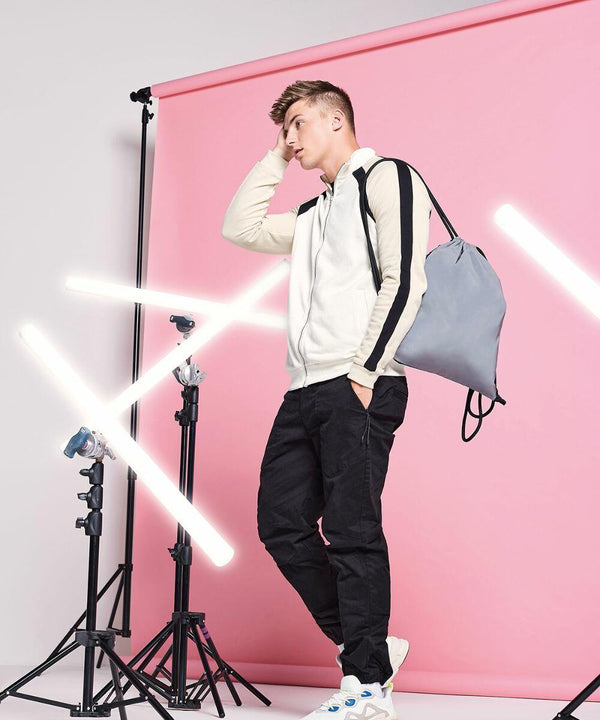 A person stands in a studio with a pink background, leaning on one leg. They wear a white and beige hoodie, black pants, and white sneakers, carrying a gray Bagbase BG137 over one shoulder. Several bright white lights and light stands are positioned around them.