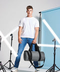 A man stands holding a gray Bagbase BG140 in front of a blue backdrop with studio lights around him. He is wearing a white T-shirt, blue jeans, and white sneakers.
