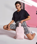 A person dressed in a black t-shirt and snake-print shorts kneels on one knee while holding a Bagbase BG153. The background features pink and white lighting setups.
