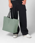 A person wearing black pinstriped trousers and white trainers is holding the Bagbase Original carryall tote bag in large sage green with black straps.