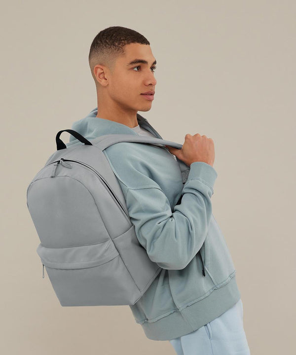 A young man in a light blue hoodie and tracksuit bottoms stands in profile, holding the straps of the Bagbase Premium Recycled Rucksack in light grey over his shoulder against a plain, neutral background.