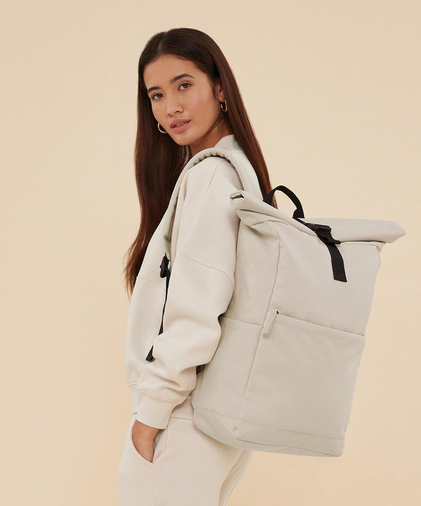 A woman with long brown hair stands against a beige background, wearing a light beige sweatshirt and joggers, carrying the Bagbase Premium Recycled Roll Top Rucksack in light beige with black straps over one shoulder.