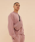 A person stands against a plain beige background, looking slightly upwards, wearing a matching light pink sweatshirt and joggers with the Bagbase Premium Recycled Cross Body Bag in pink.