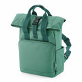 The BG18S by Bagbase is a green backpack featuring a top flap with a black buckle closure. It includes padded shoulder straps, a front zippered pocket, and side pockets. The bag is displayed upright against a plain white background.