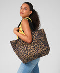 A woman with curly hair wears a yellow top and blue jeans, carrying the Bagbase Leopard print carryall tote bag over her shoulder as she stands against a plain light background facing slightly away from the camera.