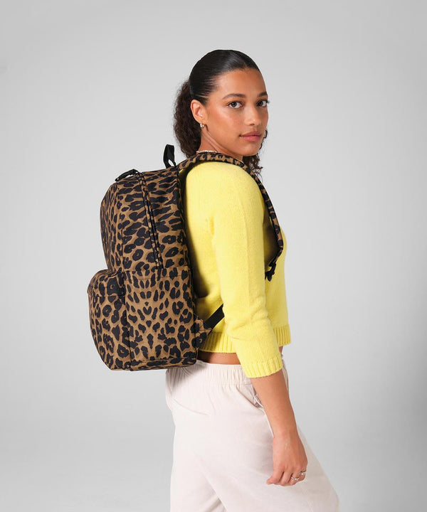 A woman with curly hair models the Bagbase Leopard print rucksack, pairing it with a yellow jumper and light trousers as she stands sideways, gazing at the camera against a plain grey backdrop.