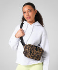 A woman with long hair tied back poses against a plain grey background, wearing a light grey hoodie and yellow trousers, accessorised with the Bagbase Leopard print cross-body bag.