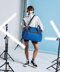 A person stands in a studio setting with a Bagbase BG200 slung over their shoulder, wearing a light grey long-sleeve top, black shorts, and black high-top sneakers.