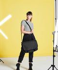 A person in a striped shirt and black jeans stands in a studio with a yellow backdrop, holding a Bagbase BG218. There are studio lights and equipment in the background.