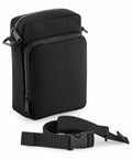 A small, black Bagbase BG241 with multiple zippered compartments is shown. The detachable and adjustable shoulder strap with a plastic buckle attachment is placed in front of the bag. The Bagbase BG241 has a minimalist design and is made of a sturdy fabric.