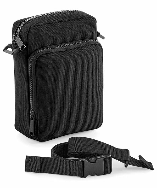 A small, black Bagbase BG241 with multiple zippered compartments is shown. The detachable and adjustable shoulder strap with a plastic buckle attachment is placed in front of the bag. The Bagbase BG241 has a minimalist design and is made of a sturdy fabric.