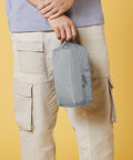 A person, cropped at the torso, is wearing light beige cargo pants and a light purple shirt, holding a Bagbase BG277 by its handle. The background is a solid yellow color.