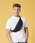 A person with short curly hair, wearing a white t-shirt and a Bagbase BG282, stands against a yellow background. They have one hand in their pocket and look directly at the camera.